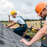Two construction workers in safety gear performing an emergency roof repair, hammering shingles on a residential roof in Madison