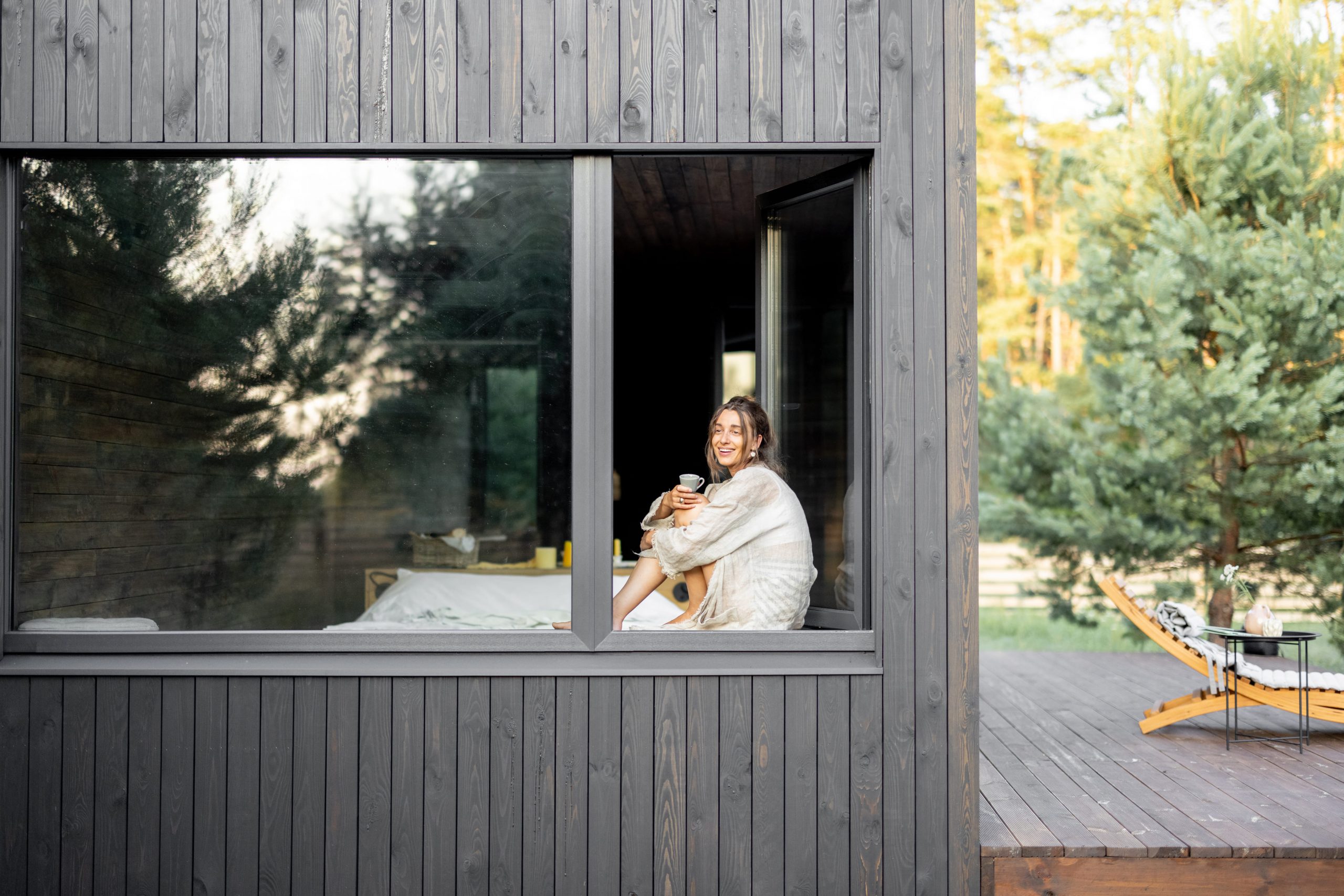 woman-sitting-by-large-window-of-modern-dark-wood-cabin-enjoying-morning-coffee